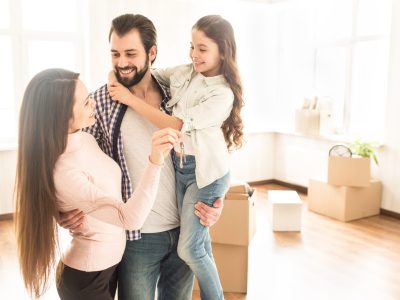 A picture of happy family standing close to each other. Man is holding his daughter in the left hand and holdin his wife with the right one. Also he is looking to his wife and smiling. It is love